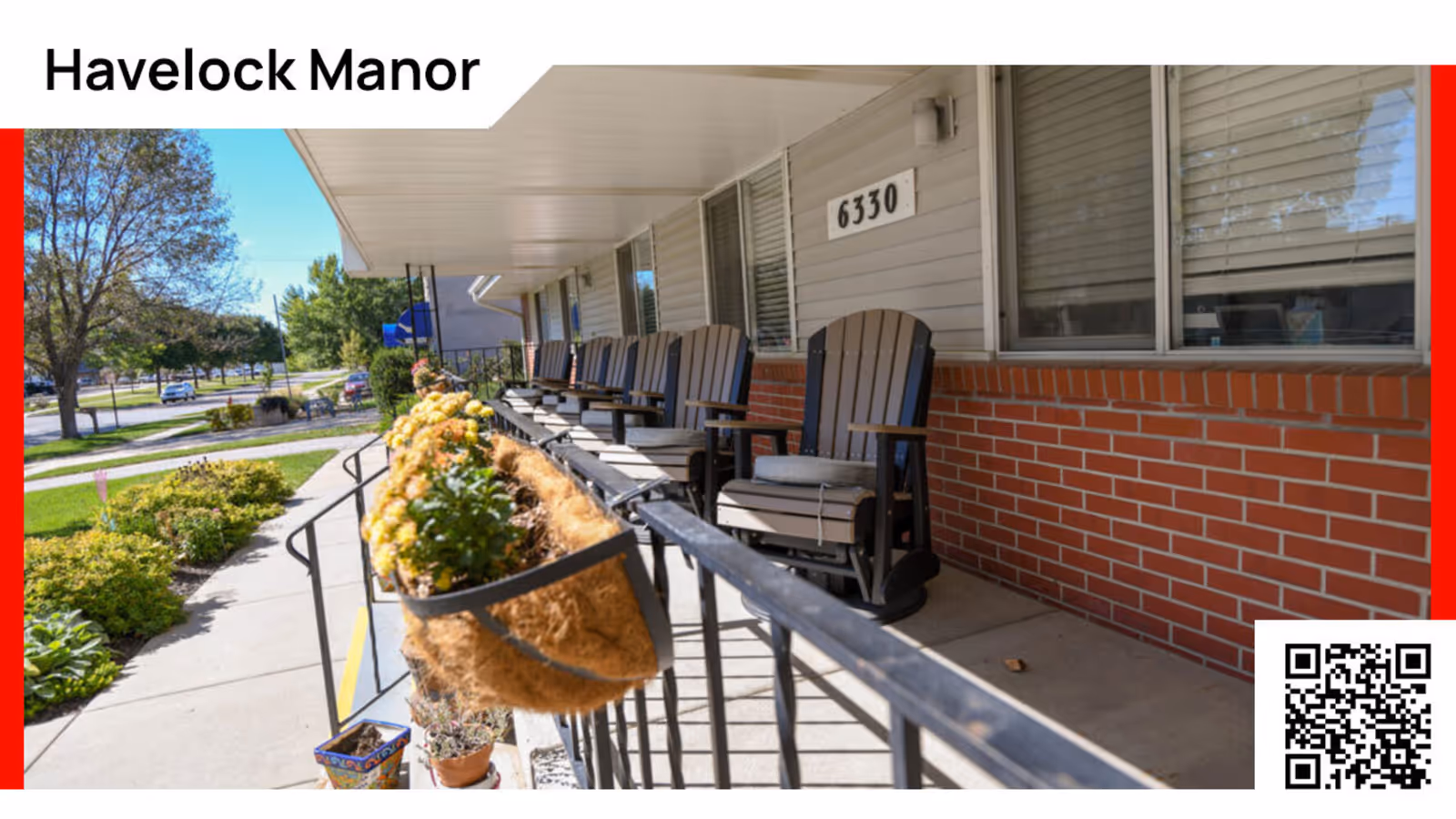 Outdoor covered porch area at Havelock Manor with several wooden rocking chairs lined up along a brick and siding wall. Flower pots with yellow flowers hang on the black metal railing, and a sidewalk and green lawn with trees are visible in the background.