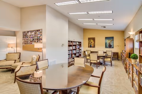 A well-lit common area with multiple wooden tables and chairs arranged for group seating. The room features bookshelves filled with books along the walls, two framed floral paintings on a yellow accent wall, and a cozy seating area with armchairs and a floor lamp. The space has a neutral color palette with beige carpeting and white walls.