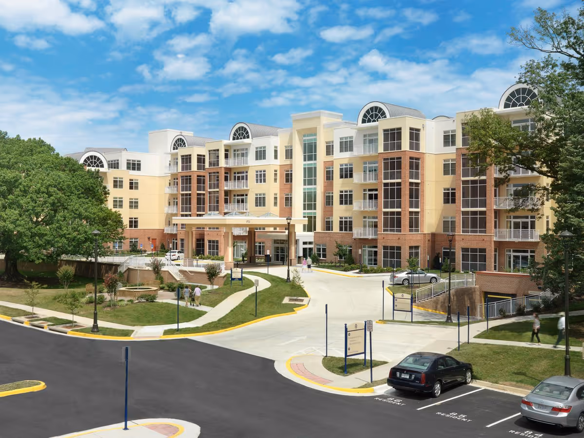 Front exterior of a multi-story retirement community building with landscaped grounds, entrance canopy, and parking lot.