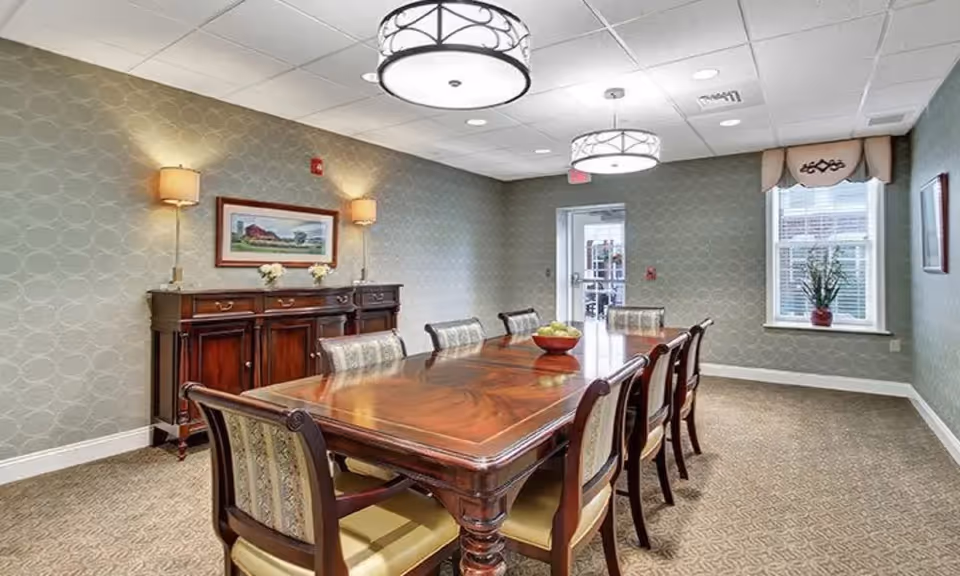 A formal dining room with a long polished wooden table, upholstered chairs, a sideboard, wall and pendant lighting, and a window.