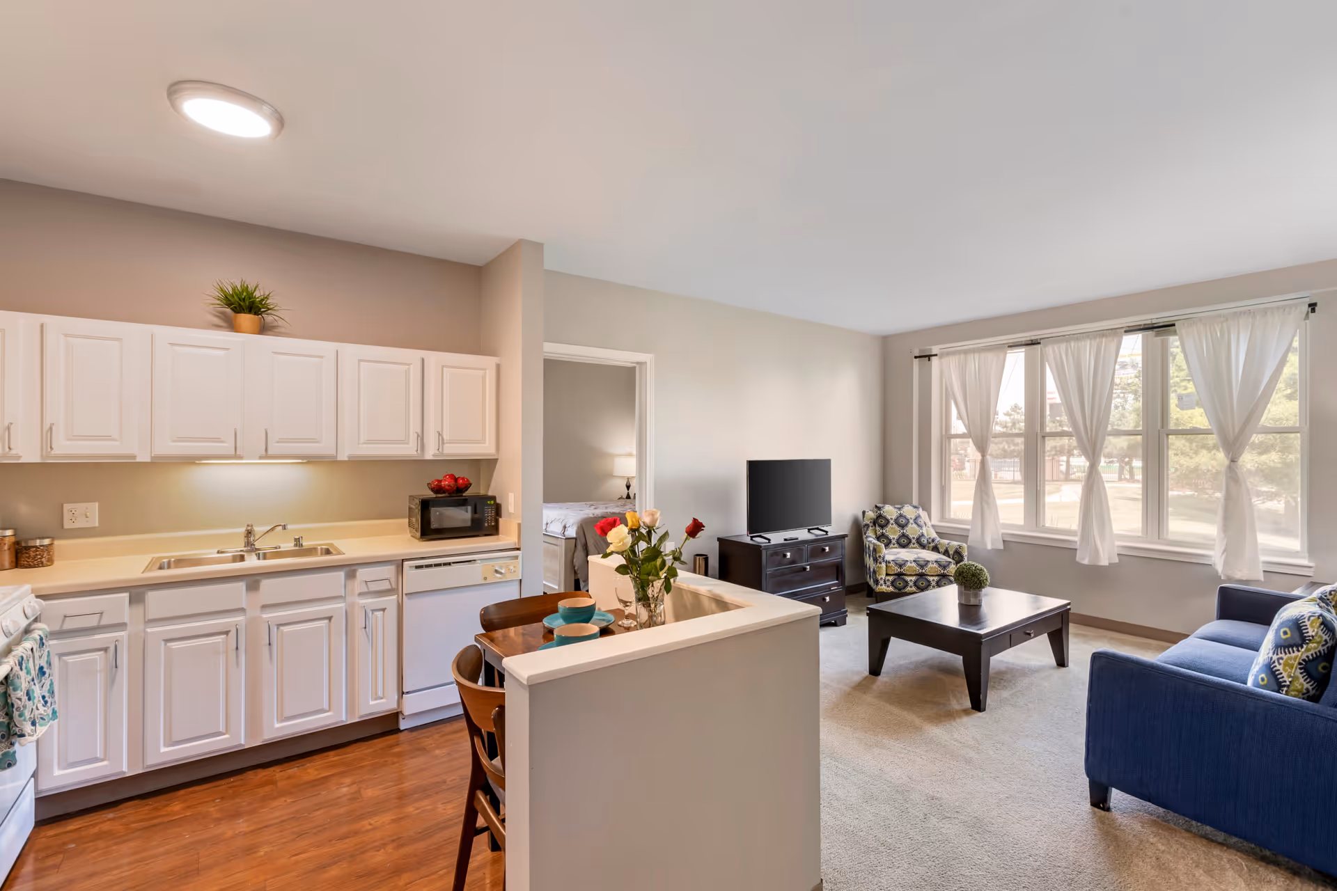 A bright and clean open-plan living space in a senior living facility featuring a kitchen with white cabinets, a sink, dishwasher, microwave, and stove. Adjacent to the kitchen is a small dining area with a wooden table set for two. The living room has a blue sofa, patterned armchair, wooden coffee table, TV on a stand, and large windows with white curtains letting in natural light.