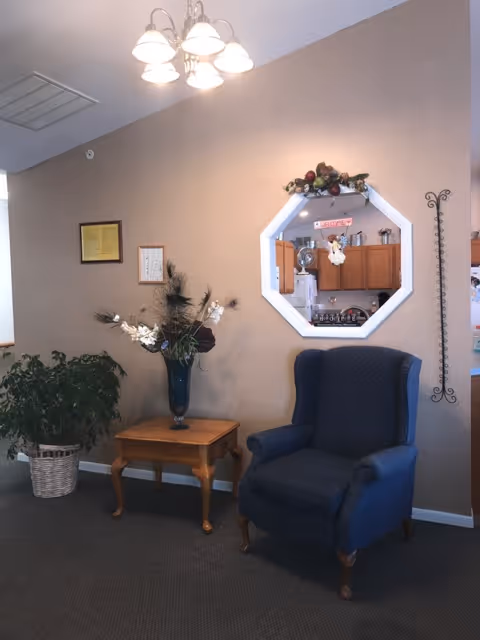 A cozy interior corner featuring a dark blue upholstered armchair next to a wooden side table with a tall vase of decorative flowers. A large leafy plant in a woven basket is placed on the floor to the left. The beige wall behind has a framed certificate and a small framed picture. An octagonal window with floral decoration on top provides a view into a kitchen area with wooden cabinets and various kitchen items. A ceiling light fixture with five lamps hangs above.
