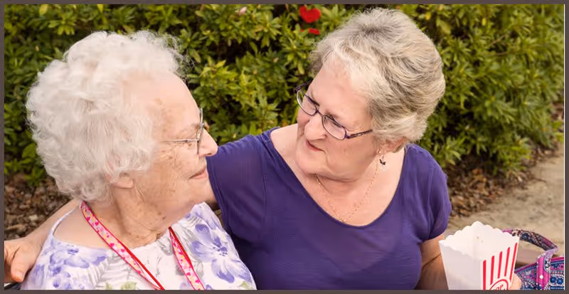 Two elderly women sitting outdoors on a bench, one wearing a purple shirt and glasses, holding a popcorn container, and the other with white curly hair and glasses, smiling at each other with greenery in the background.