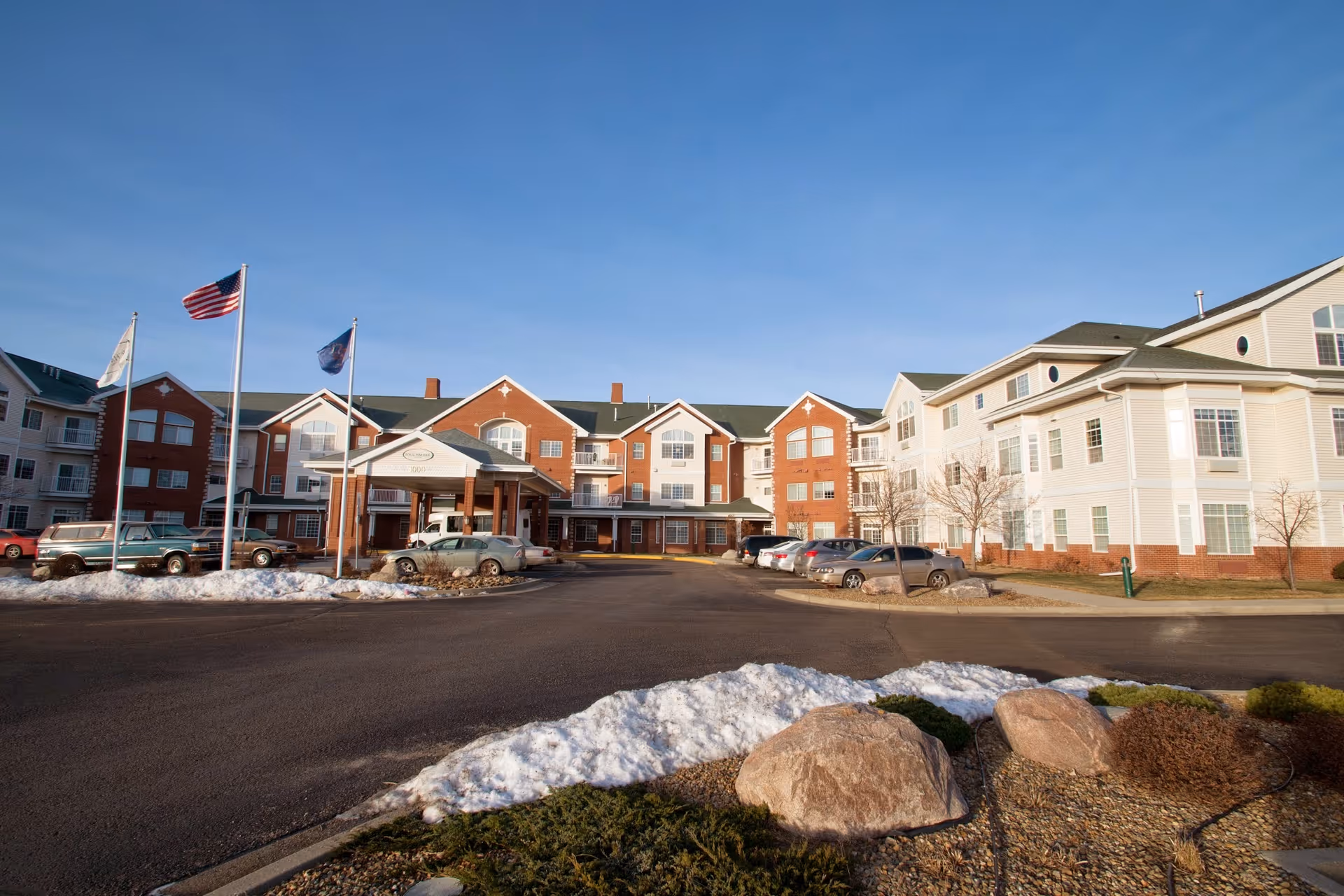 Front exterior of a multi-story senior living building with flags, a covered entrance, parked cars, and patches of snow.