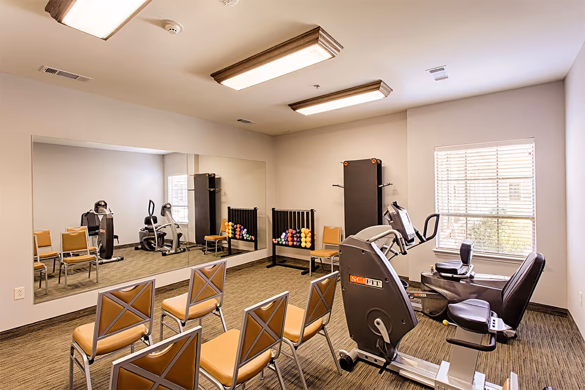 A small fitness room with exercise equipment including a recumbent bike and a stationary bike. There are several chairs arranged in rows facing a large wall mirror. A rack with colorful dumbbells and a folded exercise mat are also visible. The room has beige walls, carpeted floor, and a window with blinds allowing natural light.