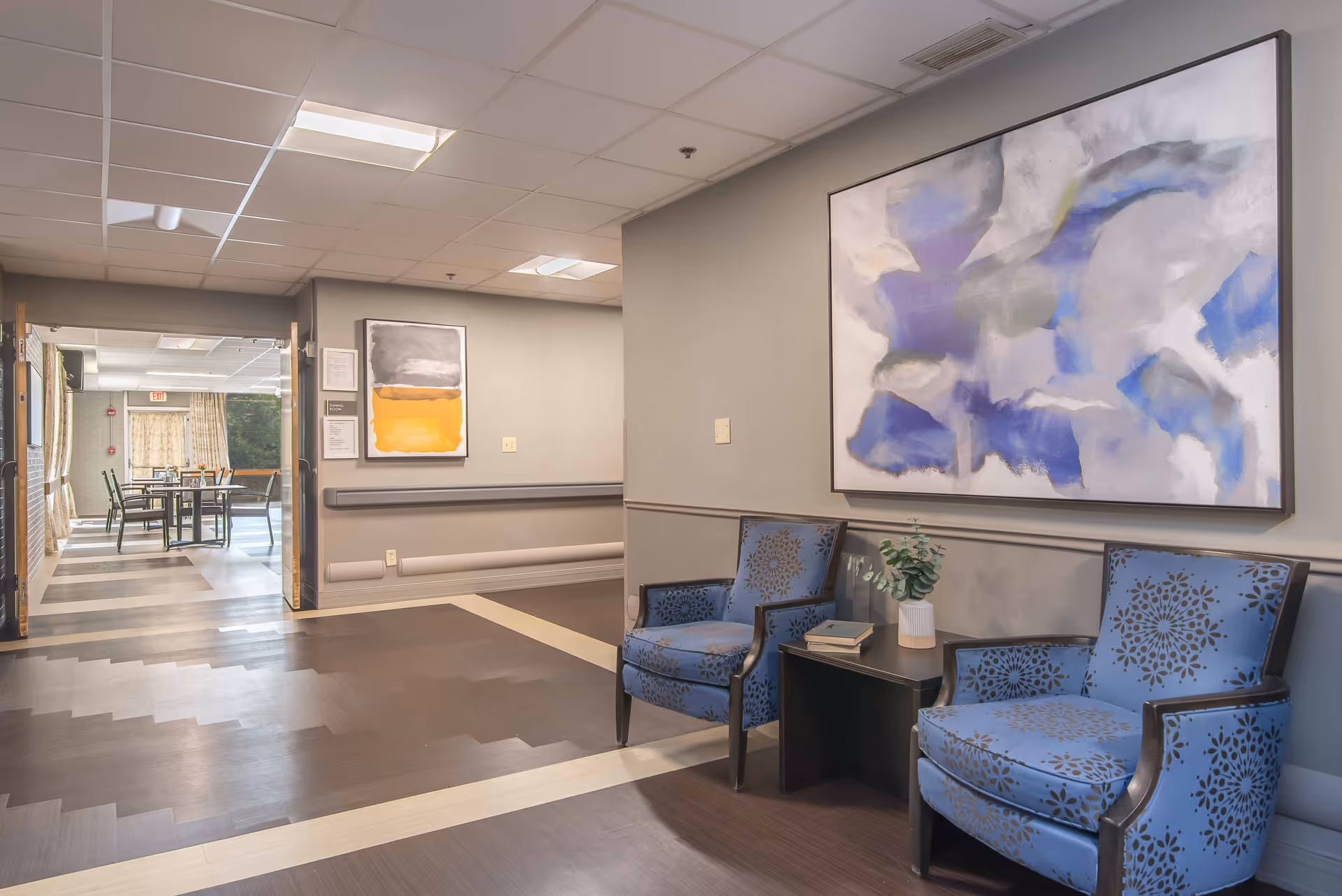 A seating area in a senior living facility with two blue patterned armchairs and a small black table between them holding a plant and a book. The walls are light gray with two abstract paintings hanging on them. The floor has a dark wood pattern with lighter wood accents. In the background, there is a dining area with tables and chairs visible through an open doorway.