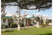 Single-story white building with a green roof, flowering trees and lawn chairs on a grassy courtyard.