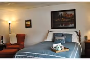 A cozy bedroom with a bed made up with blue and white bedding, several pillows, and a wooden headboard. There is a tray with a teapot and cups on the bed. To the left of the bed is a red armchair with a white throw blanket draped over it and a tall floor lamp behind it. Two framed pictures hang on the wall above the bed and chair. A wooden nightstand with a lamp and some items is on the right side of the bed.