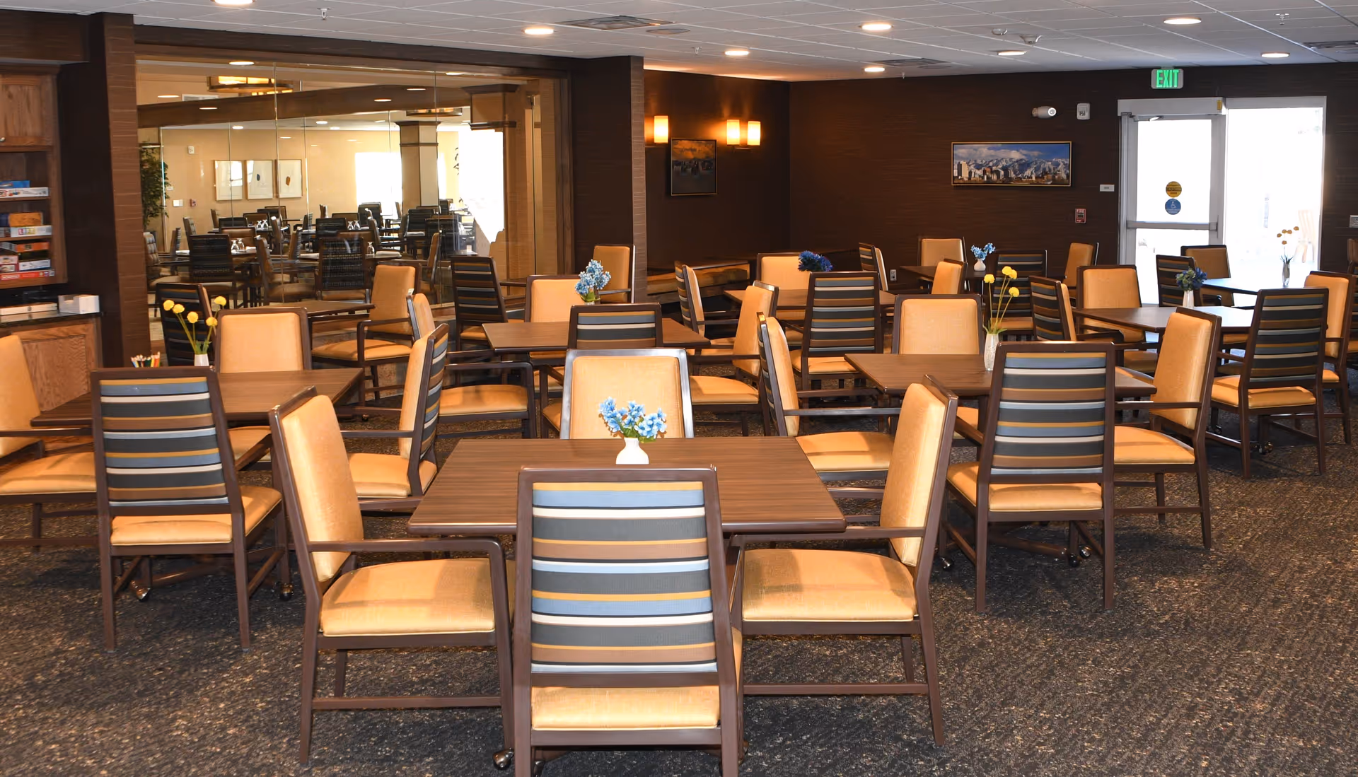 A spacious dining room with multiple wooden tables and cushioned chairs arranged neatly. Each table has a small vase with colorful flowers. The room has a carpeted floor, dark brown walls, and a large mirror reflecting the room. There is an exit door with a green exit sign above it and some framed pictures on the walls.
