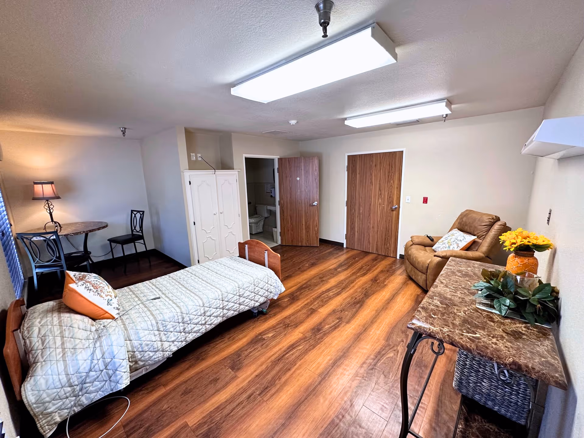 A senior living facility bedroom with a single bed covered in a beige quilt and decorative pillows. The room has wood flooring, a small round table with two chairs, a white wardrobe, a brown recliner chair, and a marble-top table with a vase of yellow flowers and a plant. Two wooden doors are visible, one slightly open showing a bathroom inside. The room is lit by ceiling lights and a table lamp.