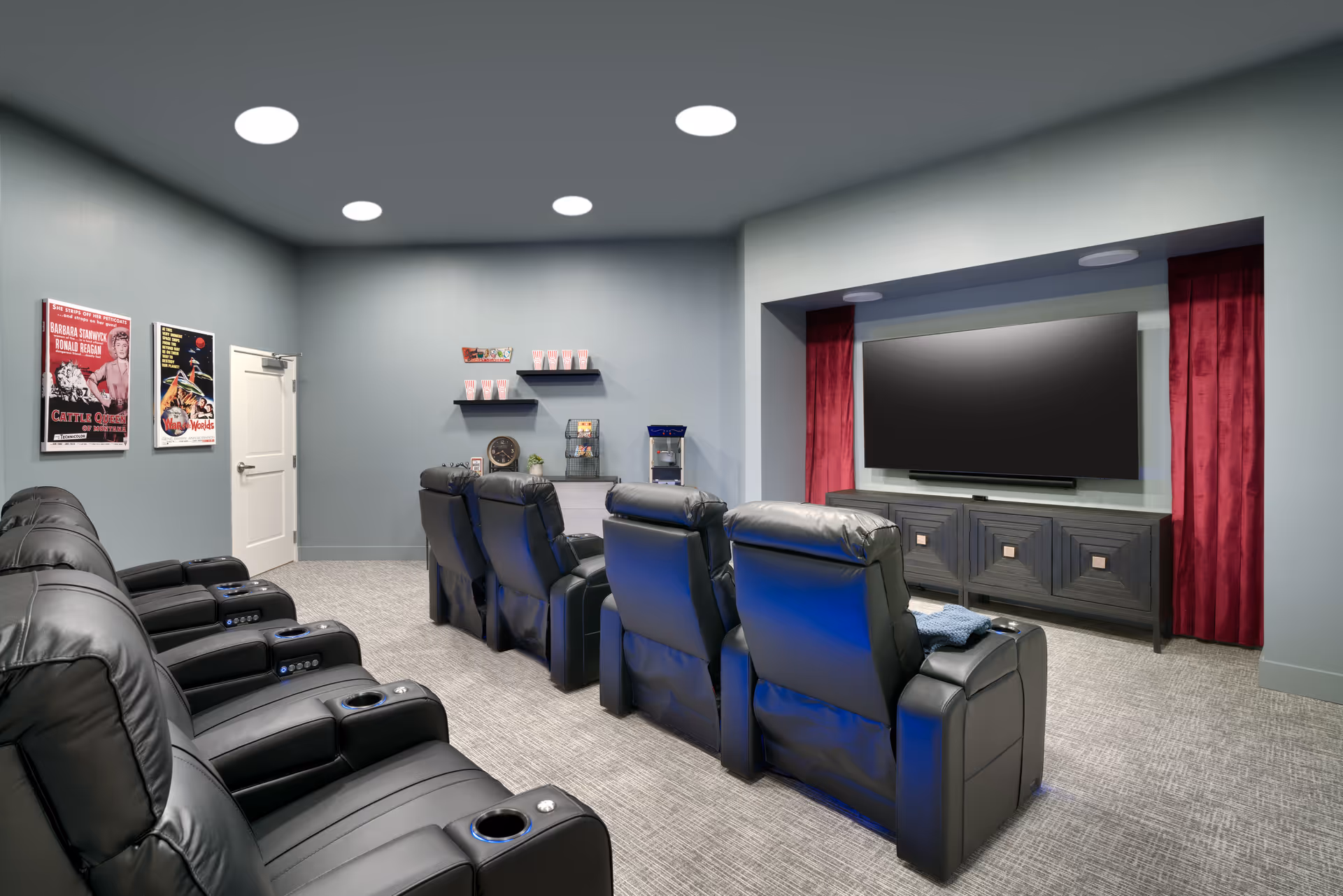 A communal home-theater room with rows of black leather recliners facing a large wall-mounted TV and a small concession area.