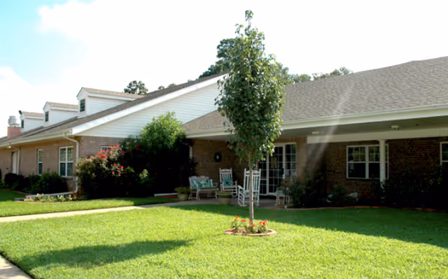 Exterior view of a single-story brick building with a well-maintained lawn, a small tree in the center, and a covered porch area with rocking chairs and potted plants.