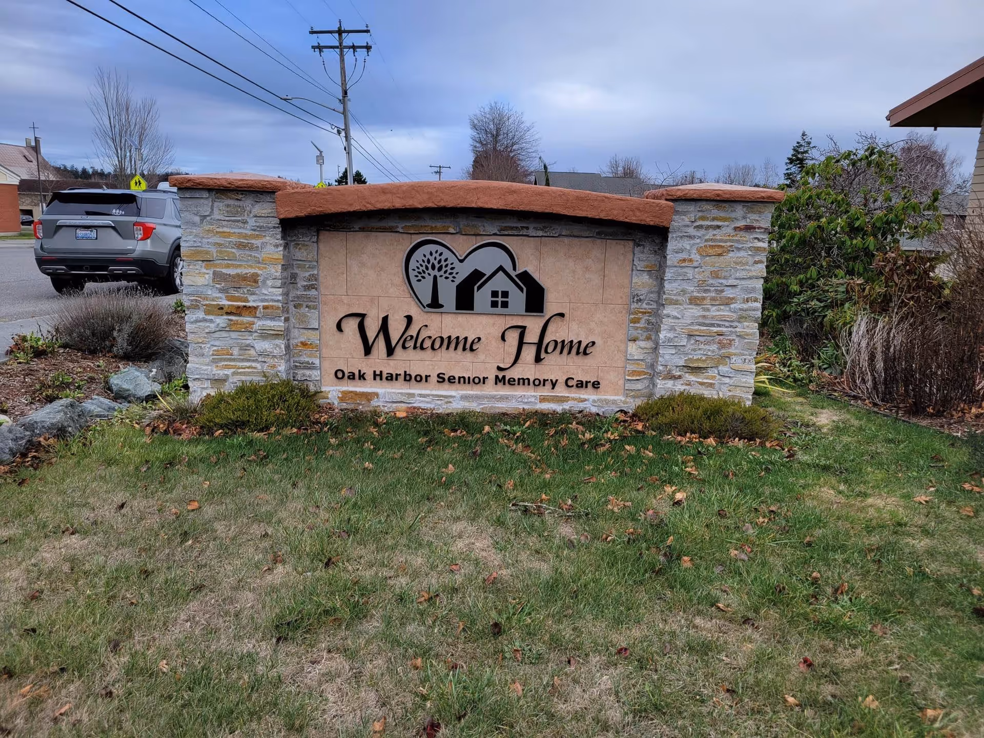 Stone and tile sign for Welcome Home Oak Harbor Senior Memory Care situated on a grassy area with bushes and a parked car visible in the background under a cloudy sky.