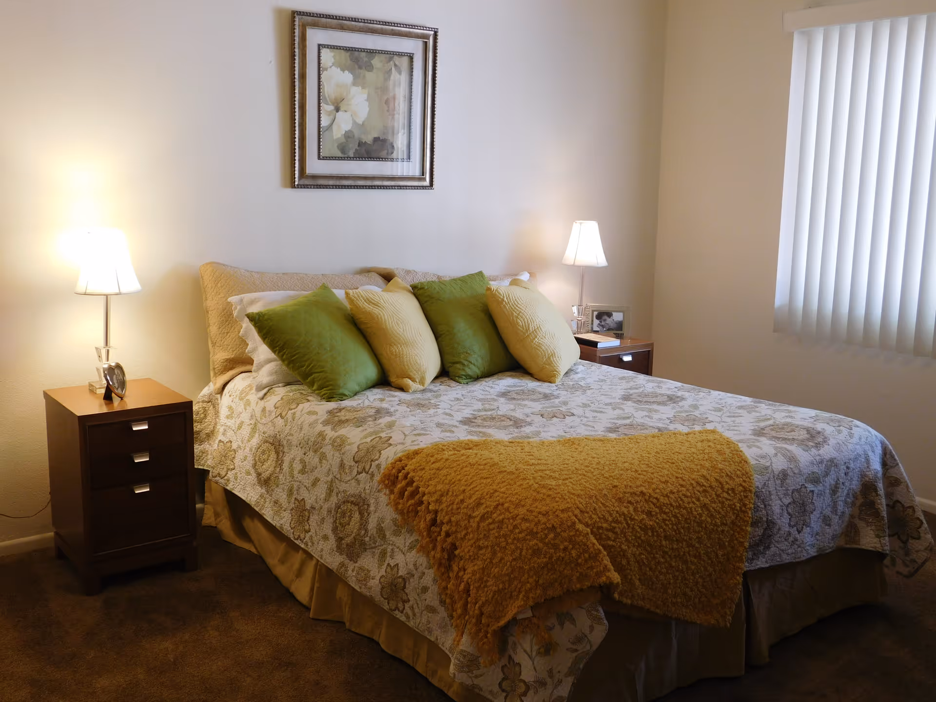 A neatly made bedroom with a patterned bedspread, green and yellow pillows, bedside tables and lamps, and framed artwork above the bed.
