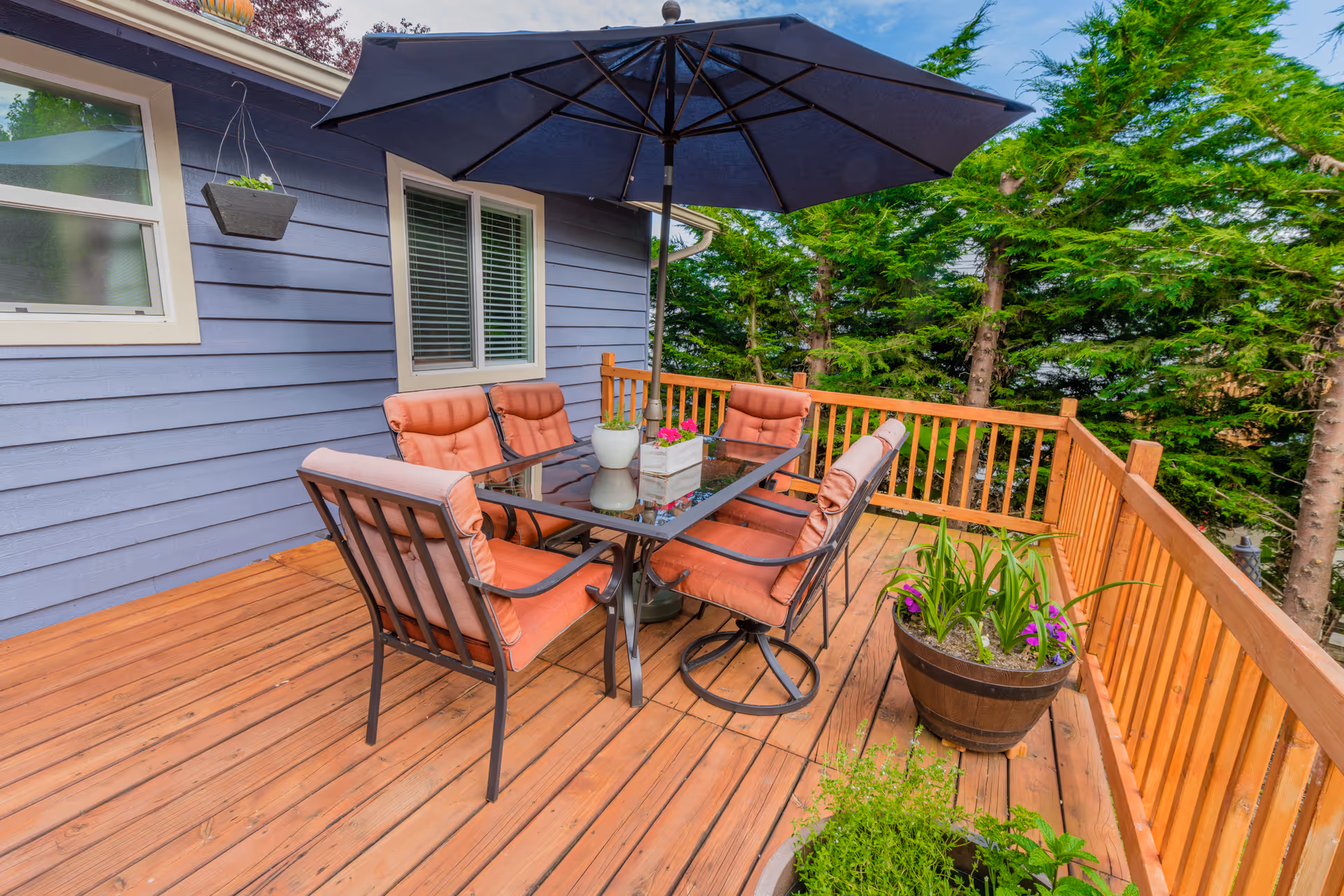 Outdoor wooden deck with a glass-top table surrounded by six cushioned chairs. A large dark blue umbrella is positioned in the center of the table. There are potted plants on the deck railing and floor, and the deck is adjacent to a blue house with windows. Trees are visible in the background.