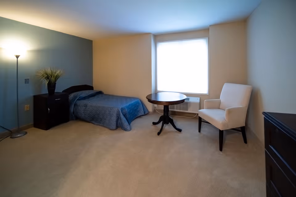 A simple bedroom with a single bed covered in a blue quilt, a dark wooden nightstand with a potted plant, a round wooden table, a white upholstered armchair, a floor lamp, and a large window letting in natural light.