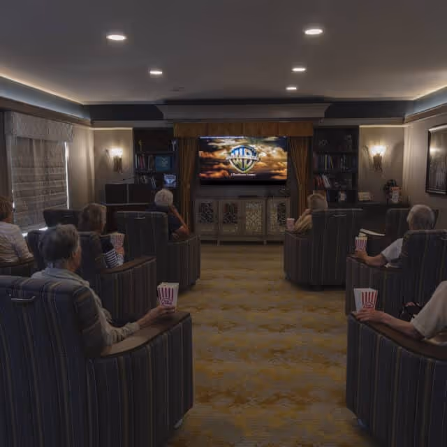 Several elderly residents sit in armchairs holding popcorn while watching a movie on a television in a dimly lit common room.