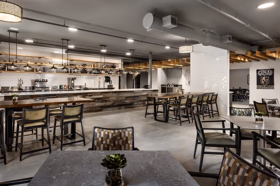 A modern dining area with multiple tables and chairs arranged neatly. The space features a long counter with a rustic wood finish and a white tiled backsplash, equipped with coffee machines and other beverage dispensers. The ceiling has exposed ductwork and industrial-style lighting fixtures. Small potted plants are placed on the tables for decoration.