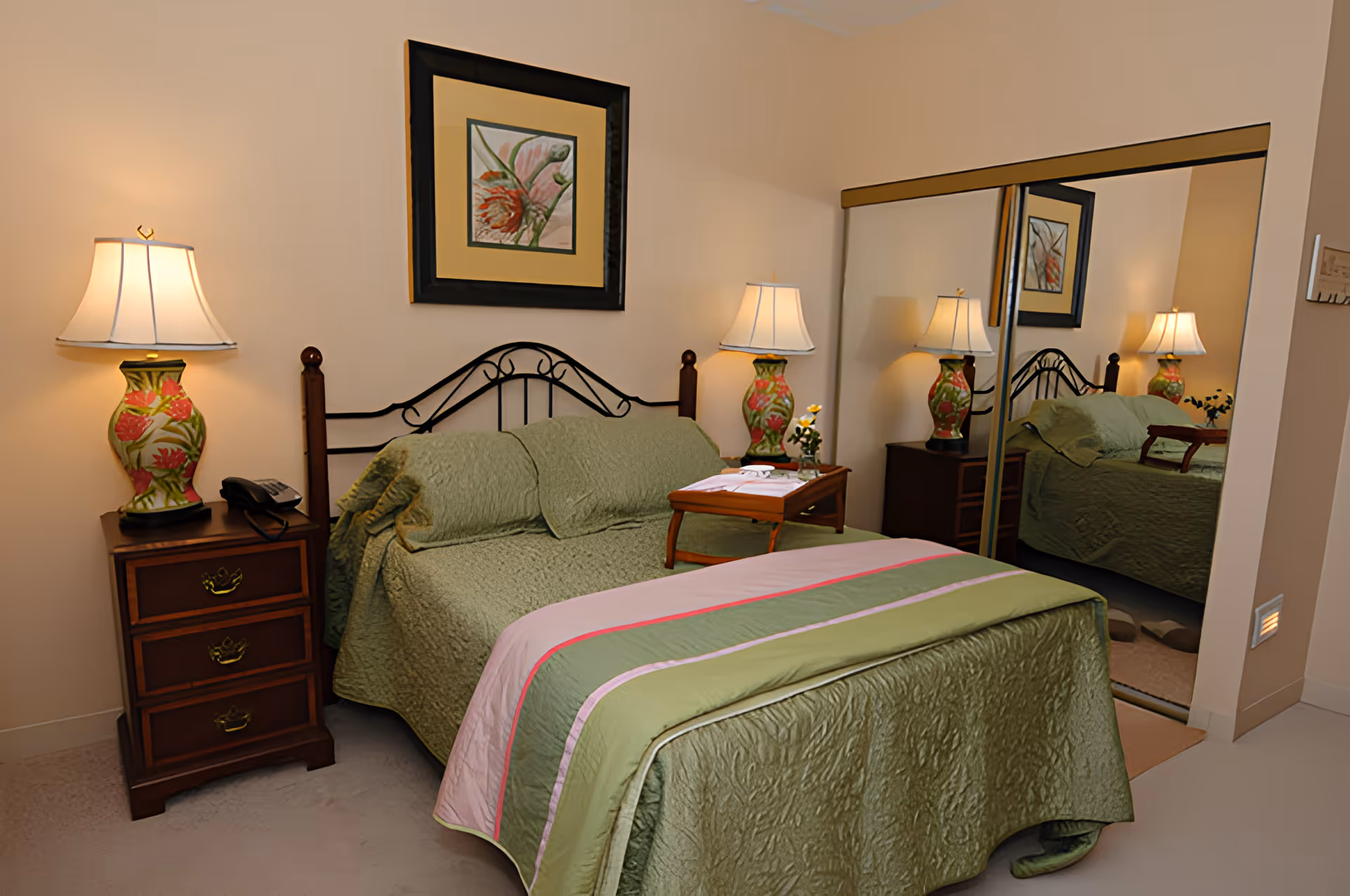 A cozy bedroom with a bed covered in green bedding and a pink and green striped blanket at the foot. There are two bedside tables, each with a decorative lamp featuring a floral design. A framed floral artwork hangs above the bed. A wooden tray with a small flower vase and a folded towel is placed on the bed. A large mirrored closet door reflects the room.