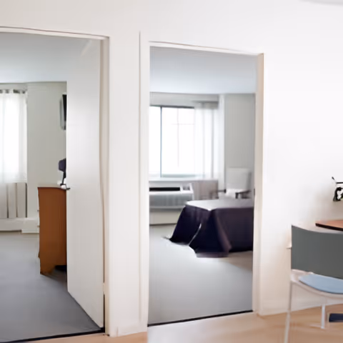 Interior view of a senior living suite with two open doorways, a bed visible through the center doorway and a small table and chair in the foreground.