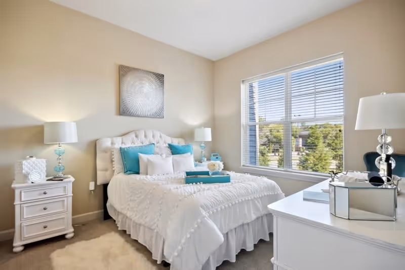 A bright and cozy bedroom with a large window letting in natural light. The room features a white tufted headboard bed with white bedding and turquoise accent pillows. There are two matching bedside tables with lamps, a white dresser with decorative items, and a soft rug on the floor.