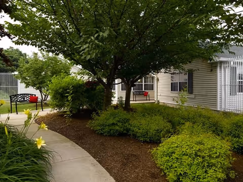 Outdoor garden area at Spring Village Stratford with a curved concrete pathway, green bushes, yellow flowers, and trees providing shade. There are two black benches with red cushions near the building with beige siding and windows.