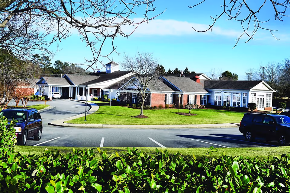 Exterior view of a single-story senior living facility building with a brick facade and white trim. The building is surrounded by a well-maintained lawn, a few leafless trees, and a parking lot with several parked cars. The sky is clear and blue.