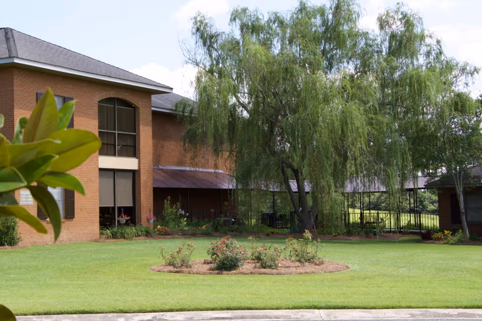 Brick building front with a manicured lawn, circular flower bed, and a large weeping willow tree.