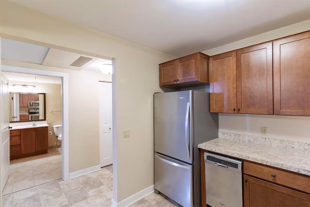 Interior view of a kitchen area with wooden cabinets, a stainless steel refrigerator, and a dishwasher. The kitchen opens into a hallway leading to a bathroom with a toilet, sink, and mirror visible.