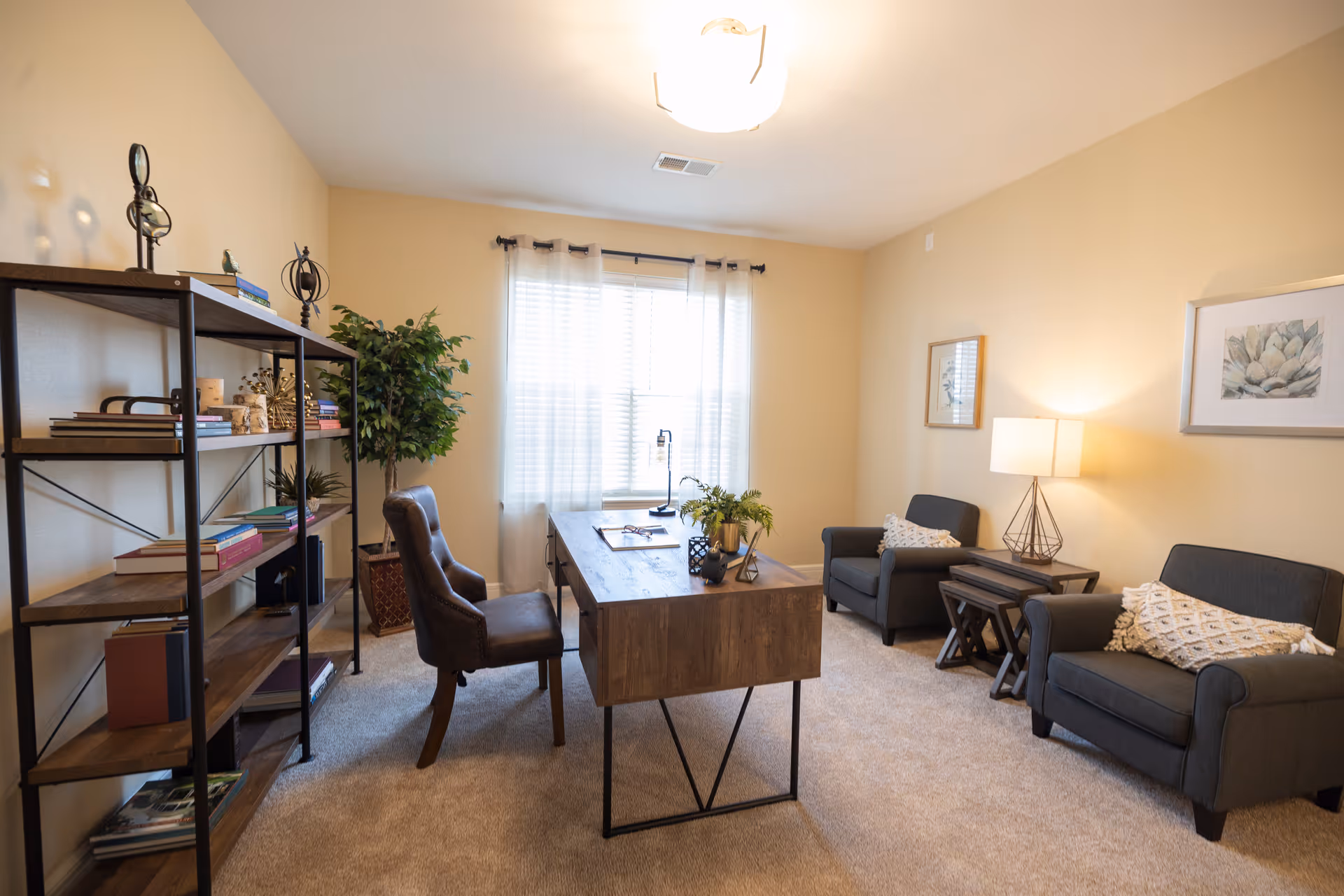 A well-lit room with beige walls and carpeted floor featuring a wooden desk with a chair, two armchairs with decorative pillows, a side table with a lamp, a tall bookshelf filled with books and decor items, and a large window with sheer curtains letting in natural light.