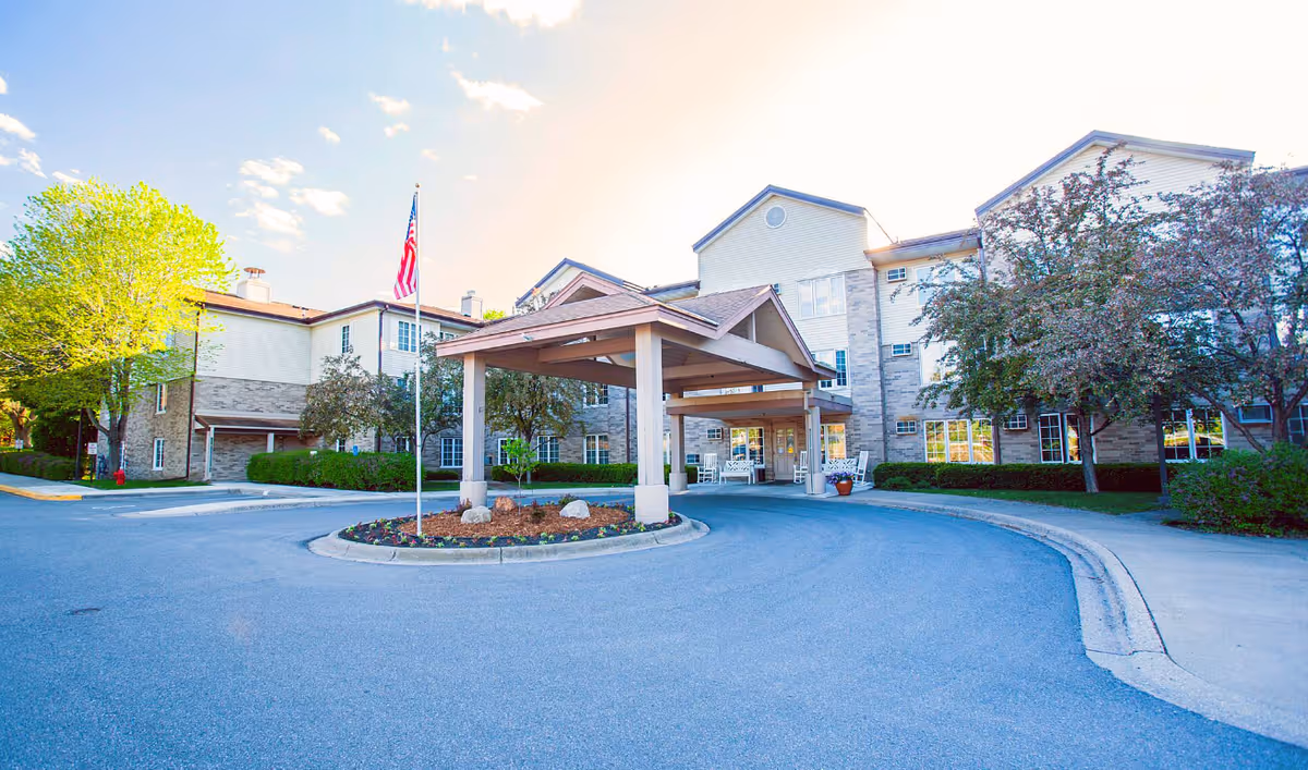 Front entrance of a multi-story senior living building with a covered porte-cochere, flagpole, circular driveway and landscaping.