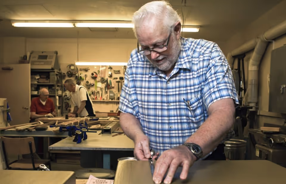 An elderly man wearing glasses and a blue plaid shirt is working with tools on a wooden project in a workshop. In the background, two other elderly men are engaged in conversation near a workbench with various tools hanging on the wall.