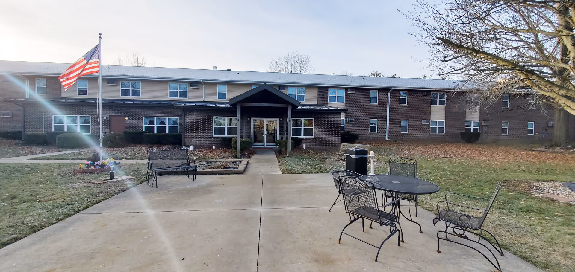 Exterior view of Cougill Senior Apartments building with a concrete patio area in the foreground featuring metal outdoor furniture including a table, chairs, and benches. An American flag is flying on a flagpole to the left, and there is a large tree on the right side of the image. The building is two stories with brick and siding exterior.
