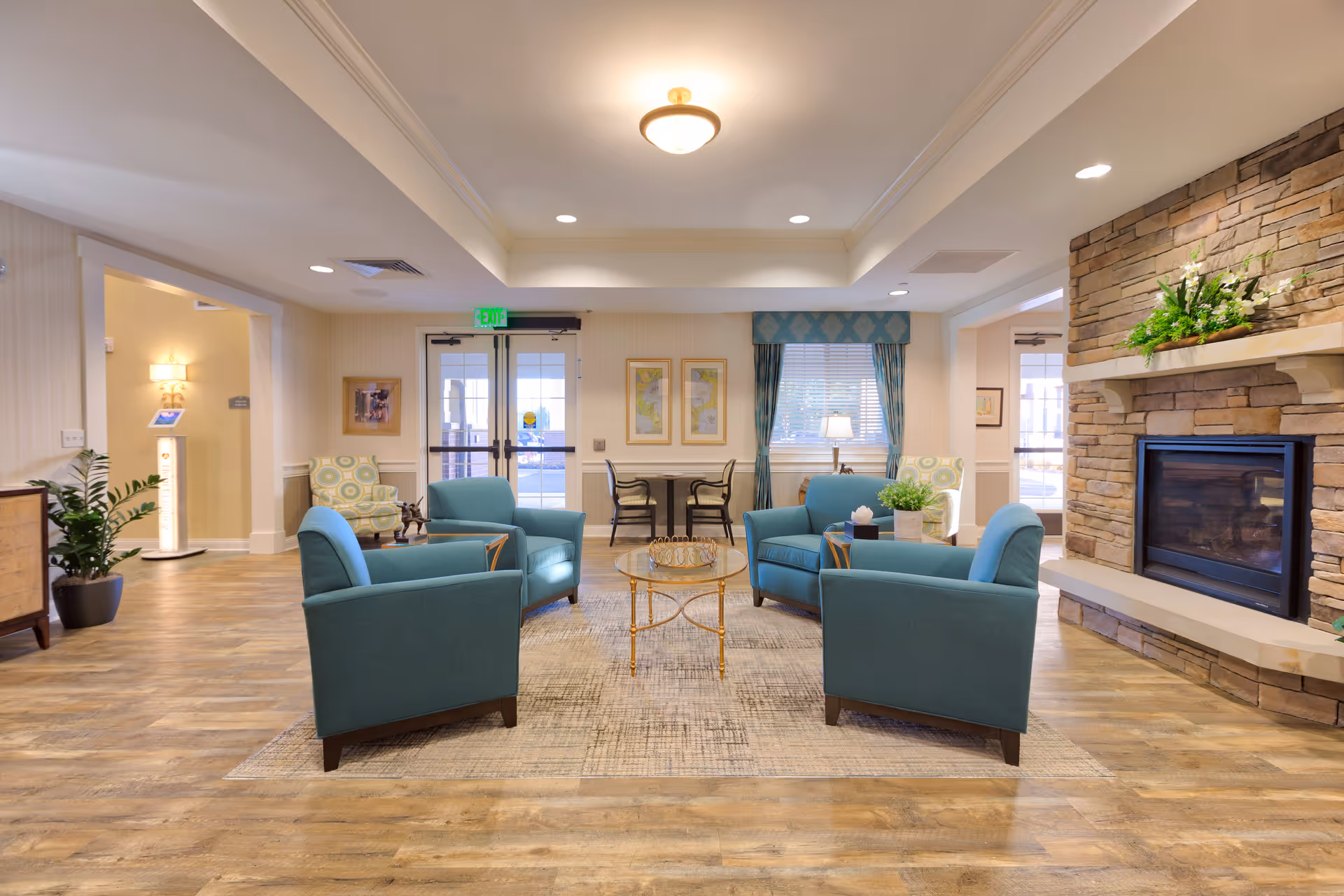 A cozy living room area in a senior living facility with four teal armchairs arranged around a glass coffee table on a patterned rug. The room features a stone fireplace with a floral arrangement on the mantel, wood flooring, and large windows with blue curtains. There are additional chairs and a small table near the windows, and double glass doors leading outside.