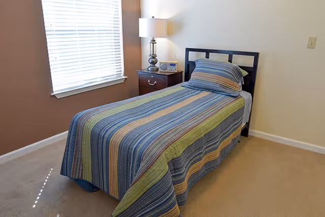 A small bedroom with a single bed covered in a striped bedspread in shades of blue, green, and beige. Next to the bed is a wooden nightstand with a lamp and a clock radio. A window with white blinds is on the left wall, letting in natural light.