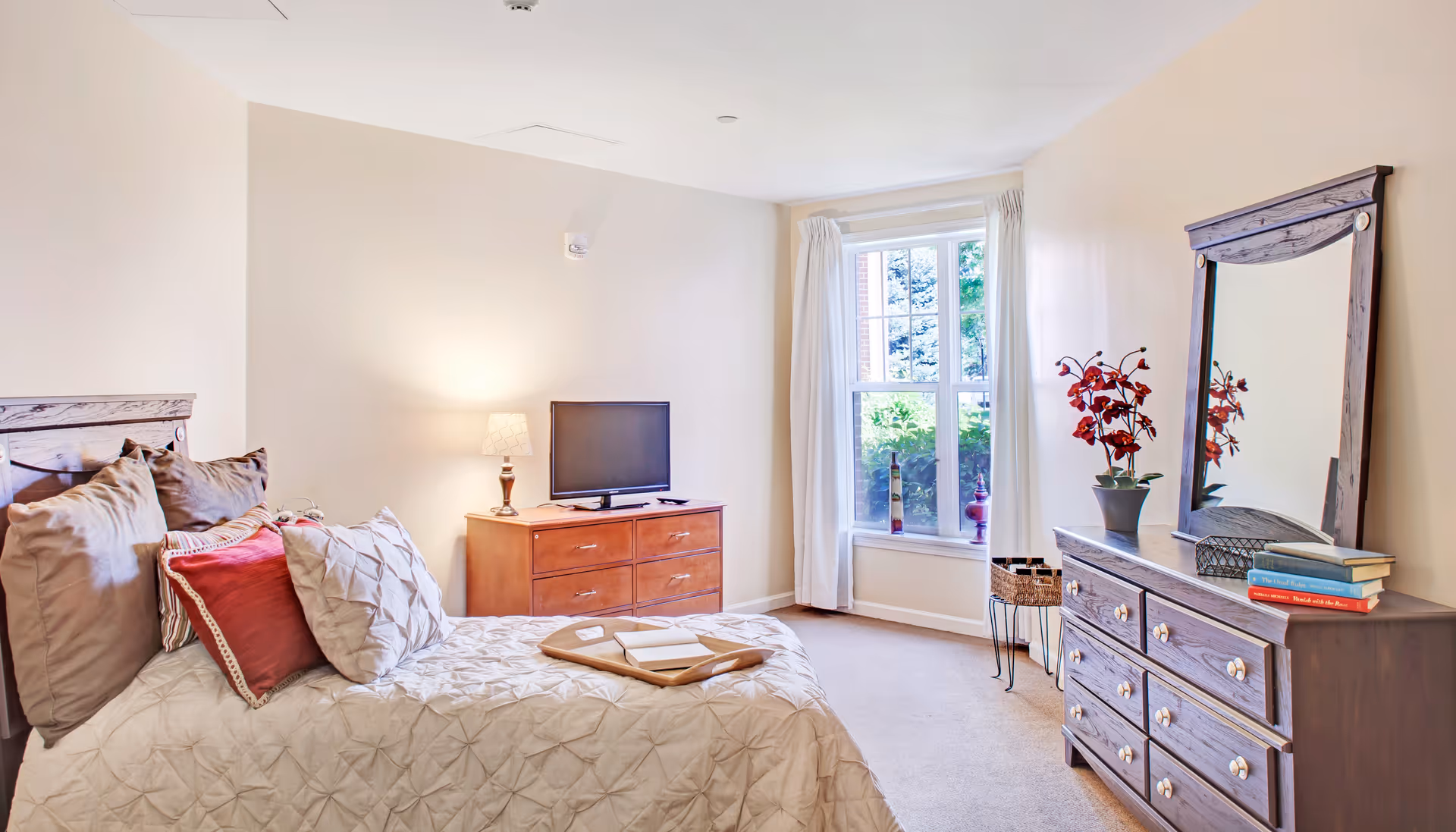 Bright, furnished bedroom with a made bed, bedside table and lamp, dresser with TV, window, and mirror.