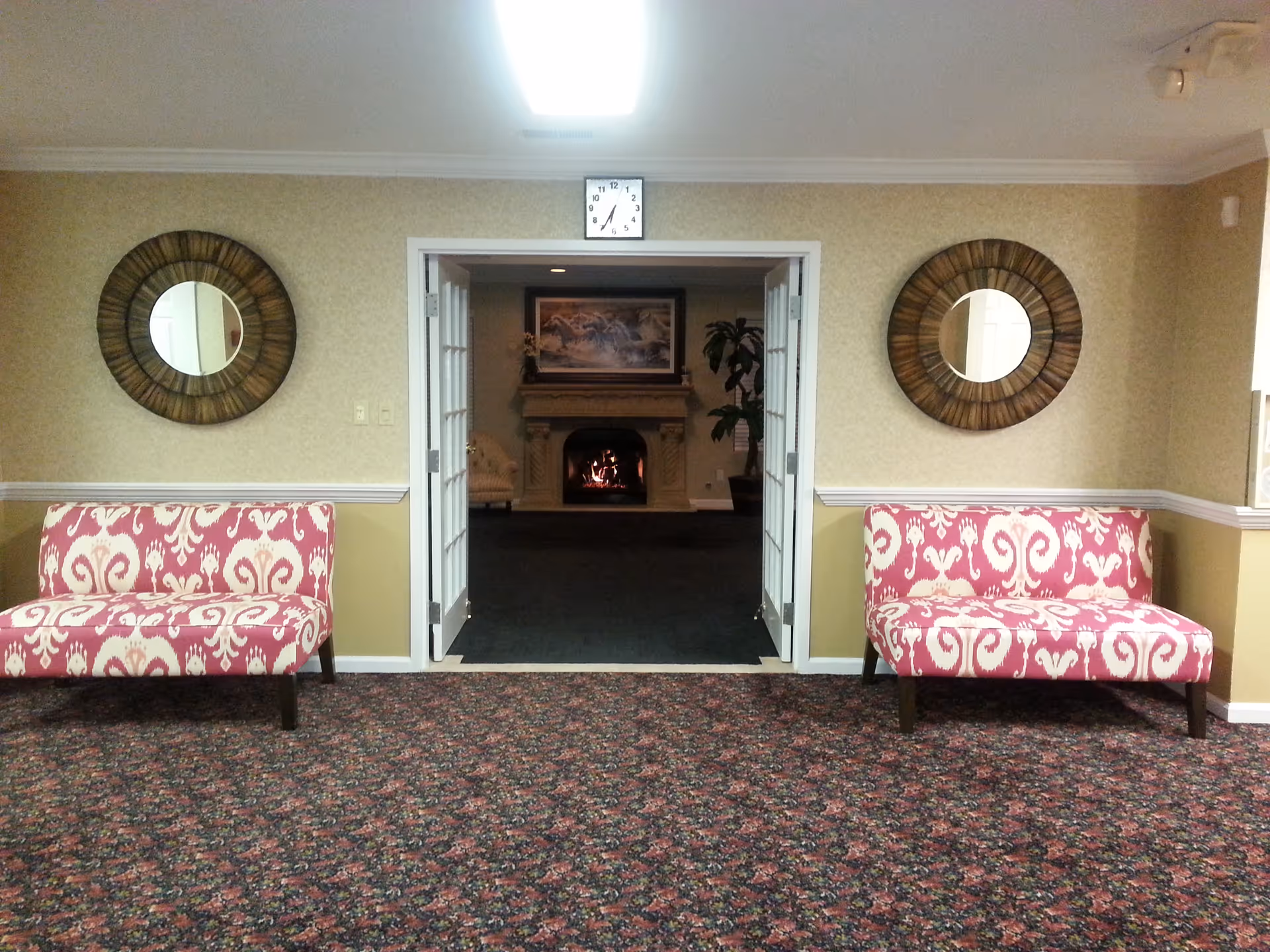 Interior view of a senior living facility lounge area with two pink patterned benches on either side of a doorway. The doorway leads to a room with a lit fireplace, a painting above it, and a potted plant. Two round mirrors hang on the walls above each bench, and a clock is mounted above the doorway. The floor is covered with a patterned carpet.