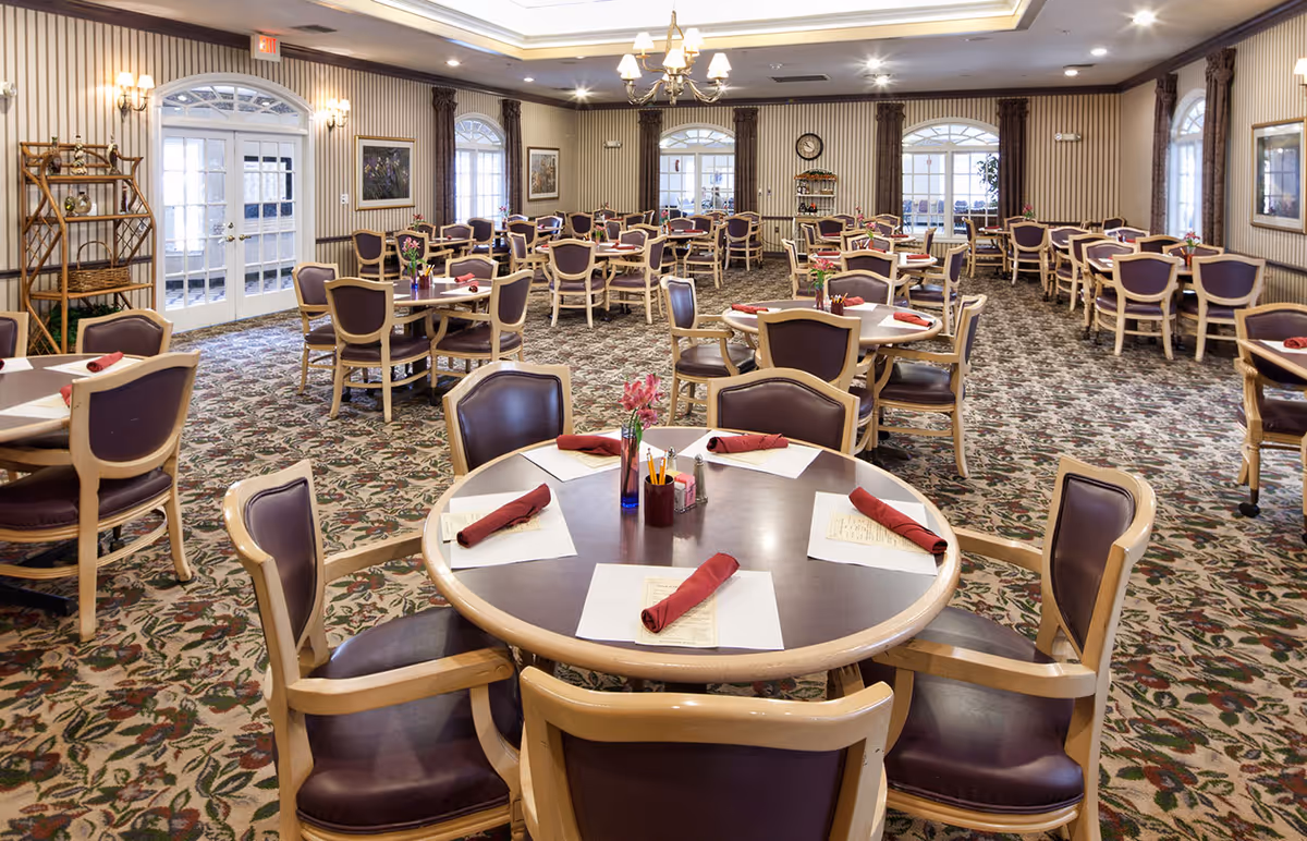 A spacious dining room with multiple round tables, each set with placemats, menus, and neatly folded red napkins. The chairs have wooden frames with dark cushioned seats and backs. The room features patterned carpet, striped wallpaper, several windows with arched tops, and a chandelier hanging from the ceiling. There are decorative elements such as framed pictures on the walls and a wooden shelving unit near the door.