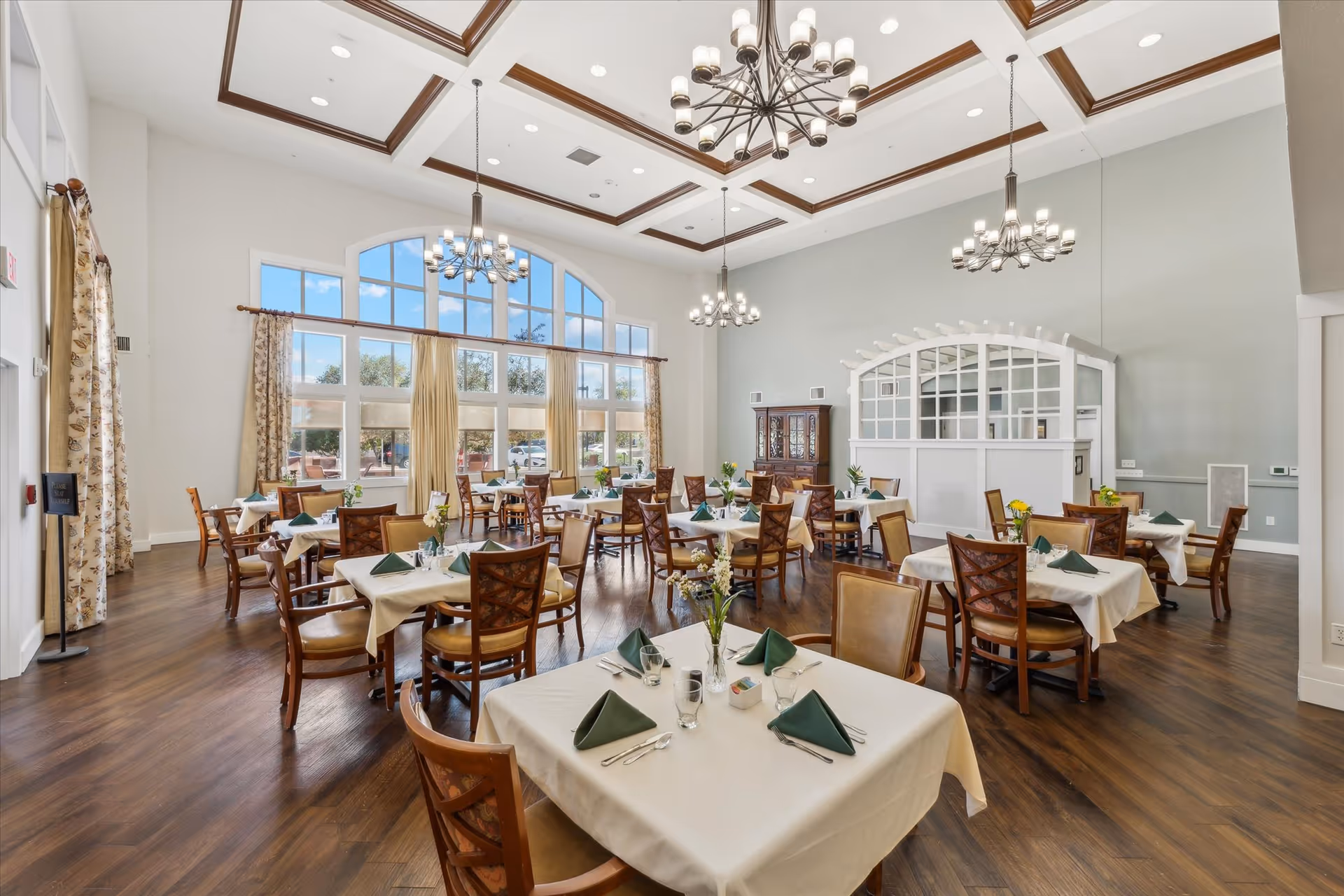 A spacious and elegant dining room with multiple tables covered in white tablecloths, each set with green folded napkins, glasses, and silverware. The room features large windows with curtains, high ceilings with decorative wooden beams, and several chandeliers providing warm lighting. The floor is wooden, and there is a cabinet and a white partition with window-like openings along one wall.
