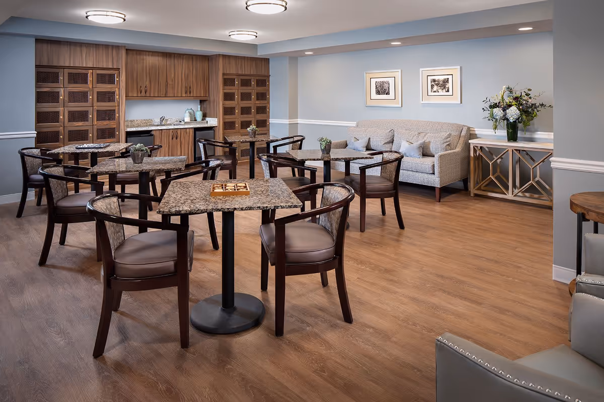A senior living facility common area with several square tables and chairs arranged on a wooden floor. The walls are painted light blue with white trim. There is a gray couch with cushions against one wall, above which hang two framed black and white pictures. A console table with a flower arrangement is positioned next to the couch. The back wall features wooden cabinetry with a granite countertop and decorative items. The room is well-lit with ceiling lights.