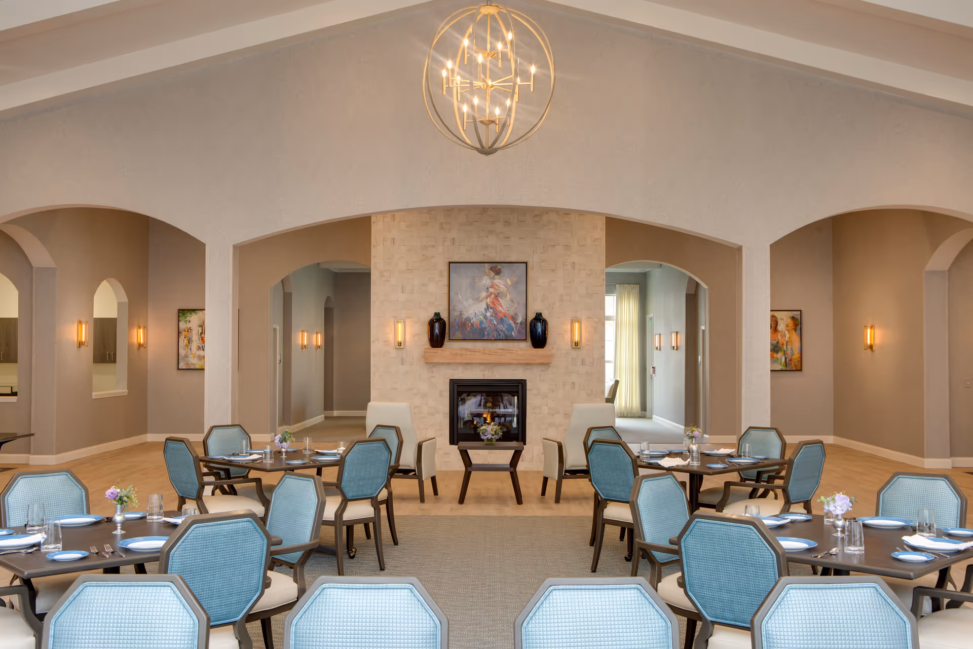A spacious dining room with multiple round tables set with plates, glasses, and silverware. The chairs have light blue upholstery and dark wooden frames. The room features a central fireplace with a painting above it and two vases on the mantel. The walls are painted in a neutral tone with soft lighting fixtures, and a modern chandelier hangs from the ceiling. There are arched doorways leading to other areas of the facility.