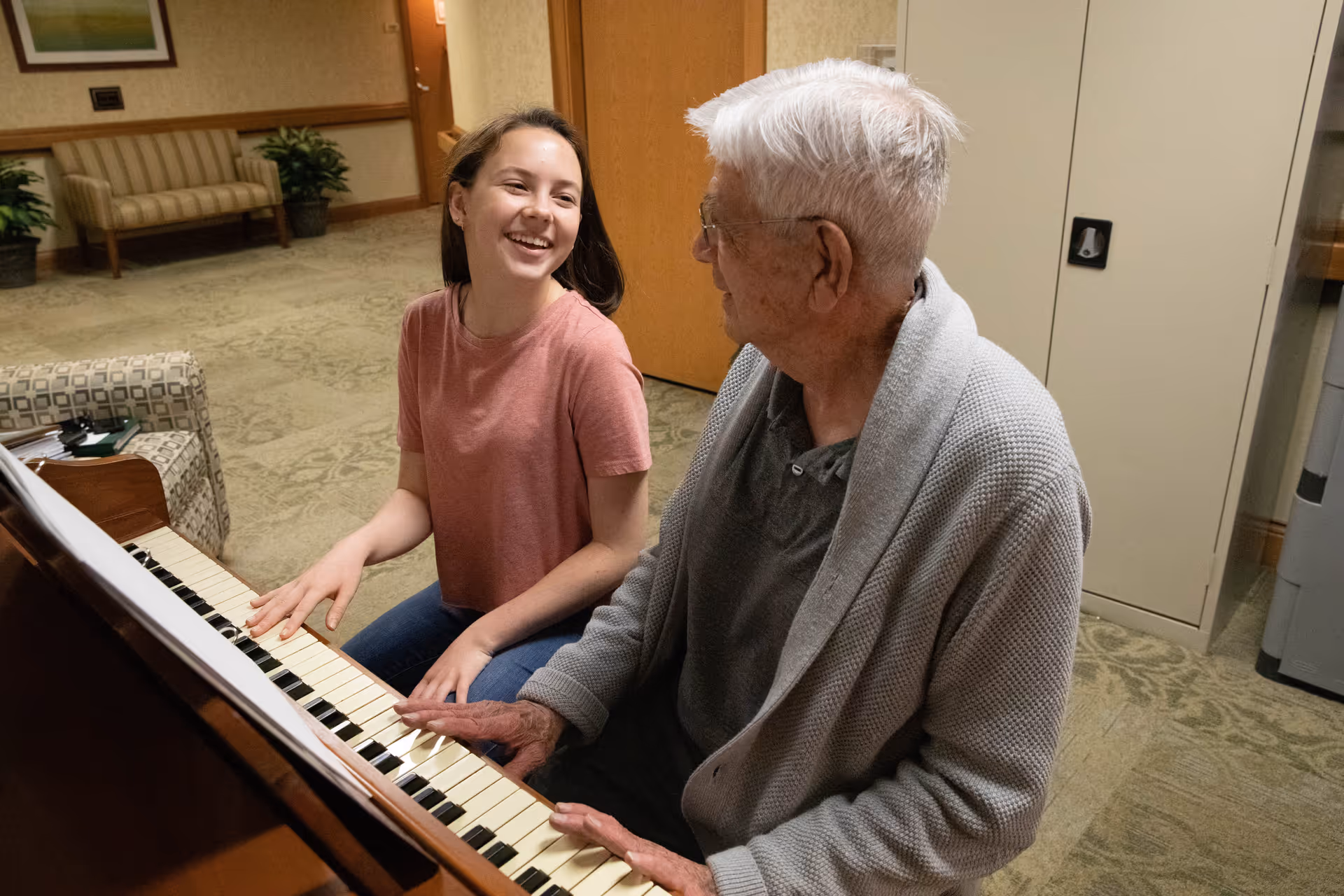 An elderly man and a young woman sitting together at a piano in a cozy room. The man is playing the piano while the woman smiles and looks at him. The room has patterned carpet, a cushioned bench, potted plants, and a wooden door in the background.