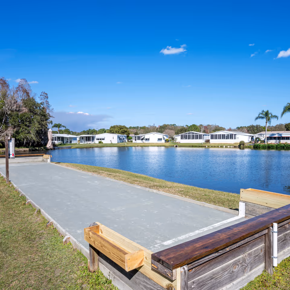 A serene outdoor scene at Cypress Lakes Village featuring a bocce ball court in the foreground, a calm pond in the middle, and several white residential buildings with screened porches in the background under a clear blue sky.