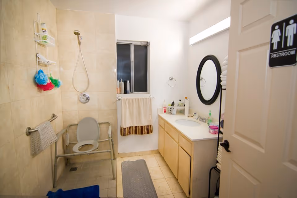 A bathroom with beige tiled walls and floor, featuring a shower area with a handheld showerhead and a shower chair with armrests. There are colorful bath sponges hanging on a wall rack, a towel hanging on a bar, and various toiletries on the windowsill and countertop. A round mirror is mounted above the sink on a white vanity with wooden cabinets. The door has a restroom sign indicating it is a unisex bathroom.