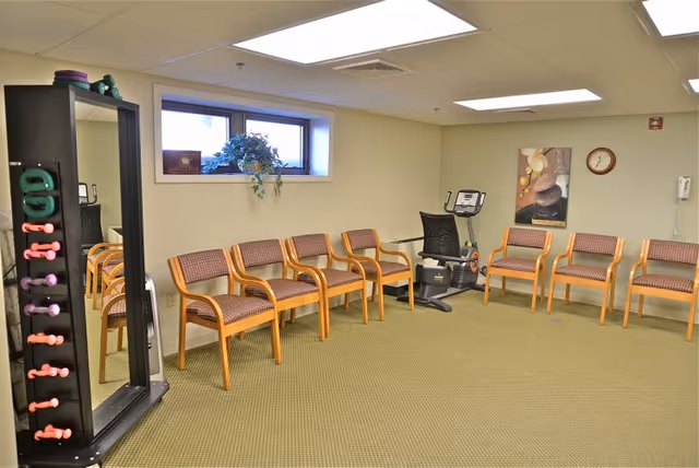 A small exercise room with a row of wooden chairs with patterned cushions arranged along two walls. There is a stationary exercise bike in the corner, a tall mirror with colorful dumbbells on its side, a small window with a plant on the sill, a wall clock, and a framed picture on the wall.