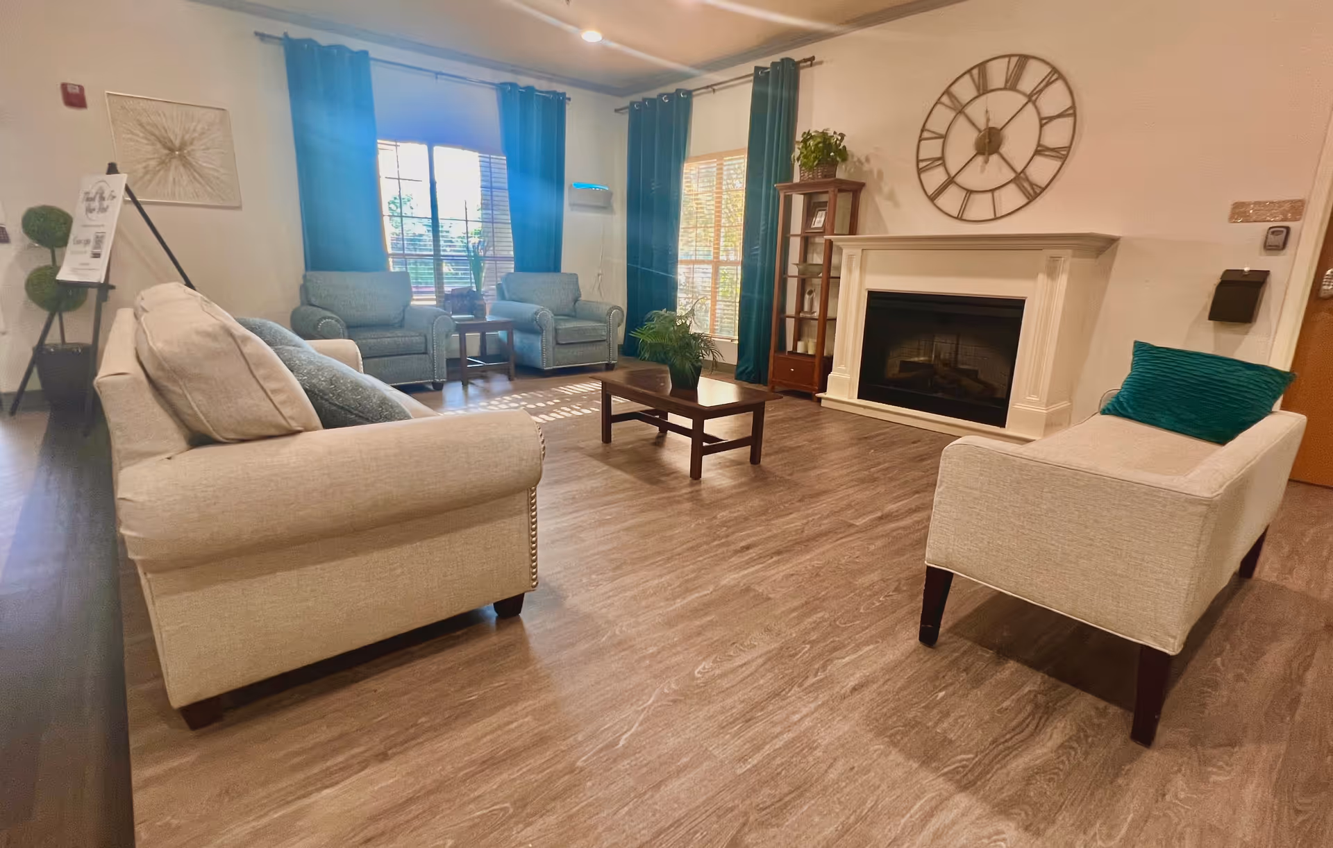 A cozy living room with beige sofas, two armchairs, a wooden coffee table, and a fireplace with a large round clock above it. The room has large windows with dark green curtains, wooden flooring, and a tall wooden shelf with plants and decor items.