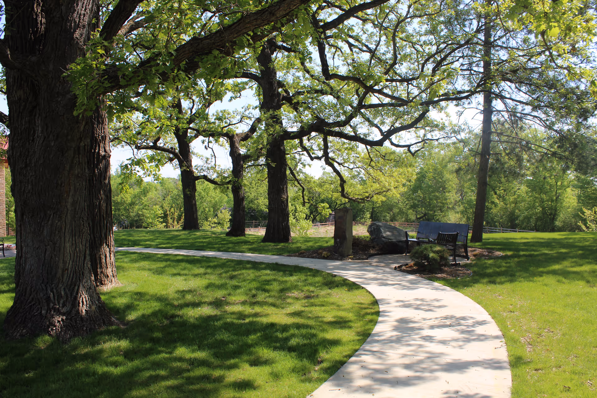A curved concrete pathway winds through a green grassy area with large trees providing shade. There are benches and a stone monument along the path, surrounded by lush greenery under a clear sky.