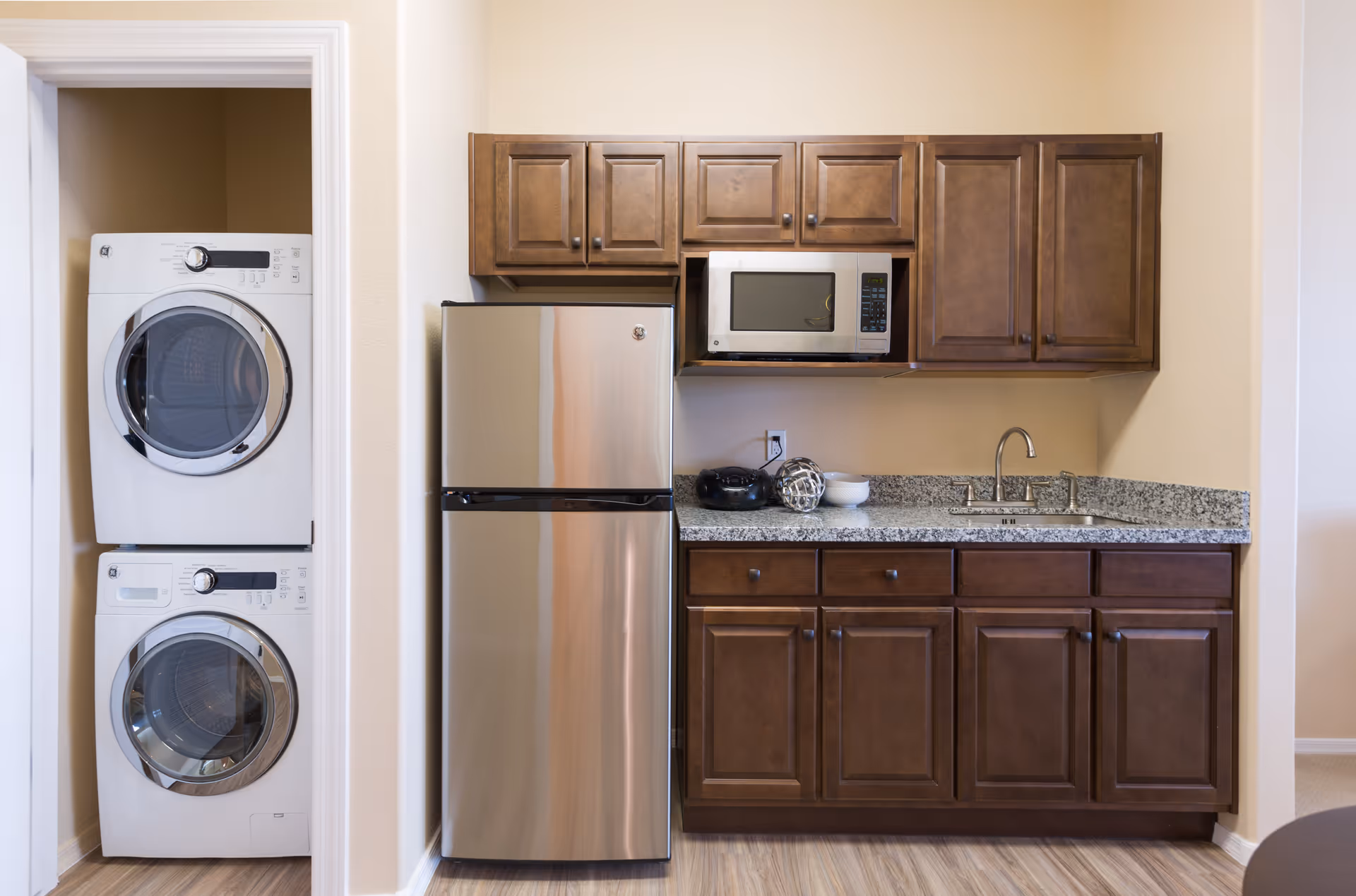 A compact kitchen area with dark wooden cabinets, a stainless steel refrigerator, a microwave oven mounted above the counter, and a granite countertop with a sink. To the left, there is a small laundry nook with a stacked washer and dryer.