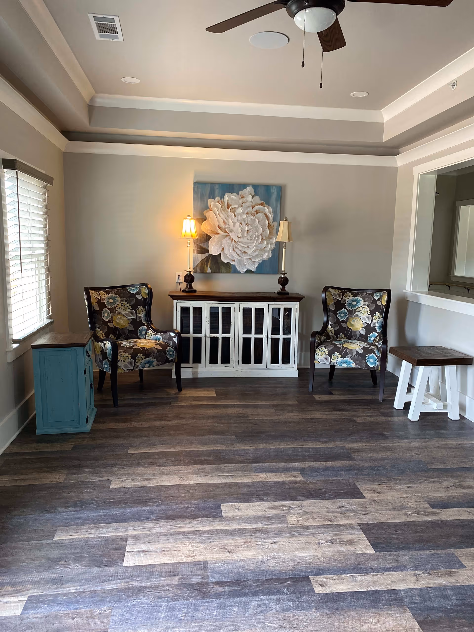 A cozy sitting area with two floral upholstered armchairs placed on either side of a white cabinet with glass-paneled doors. Two table lamps with warm light are on top of the cabinet, and a large floral painting hangs on the wall above it. The room has wood flooring, a ceiling fan, and a window with blinds on the left side. There is a small turquoise side table next to the left chair and a small wooden table with white legs next to the right chair.