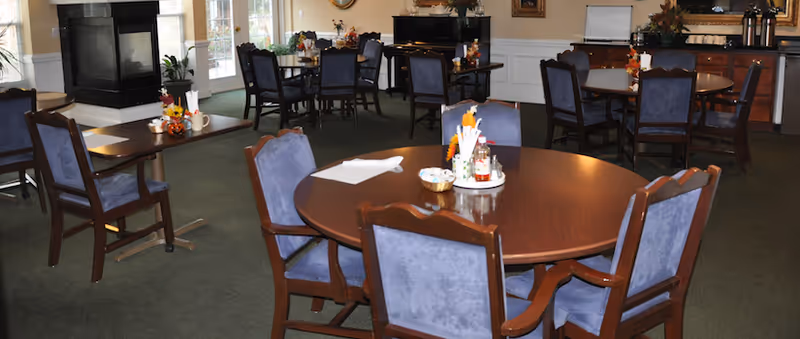 Interior view of a dining room in a retirement community with multiple round wooden tables surrounded by blue cushioned chairs. Tables have small decorative centerpieces and condiments. The room has large windows, a fireplace, and a buffet area with cabinets and a coffee station in the background.